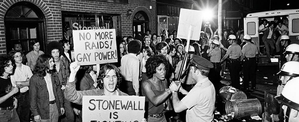 AI-generated black and white illustration depicting protesters outside the Stonewall Inn on Christopher Street in Greenwich Village, New York, during the 1969 Stonewall uprising. Crowd holds signs reading "No More Raids, Gay Power" and "Stonewall Is Fighting Back" while confronting police officers in riot gear. Note: This is an AI-generated illustration, not a historical photograph.