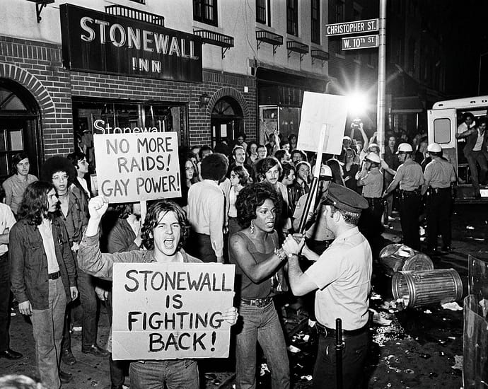 AI-generated black and white illustration depicting protesters outside the Stonewall Inn on Christopher Street in Greenwich Village, New York, during the 1969 Stonewall uprising. Crowd holds signs reading "No More Raids, Gay Power" and "Stonewall Is Fighting Back" while confronting police officers in riot gear. Note: This is an AI-generated illustration, not a historical photograph.